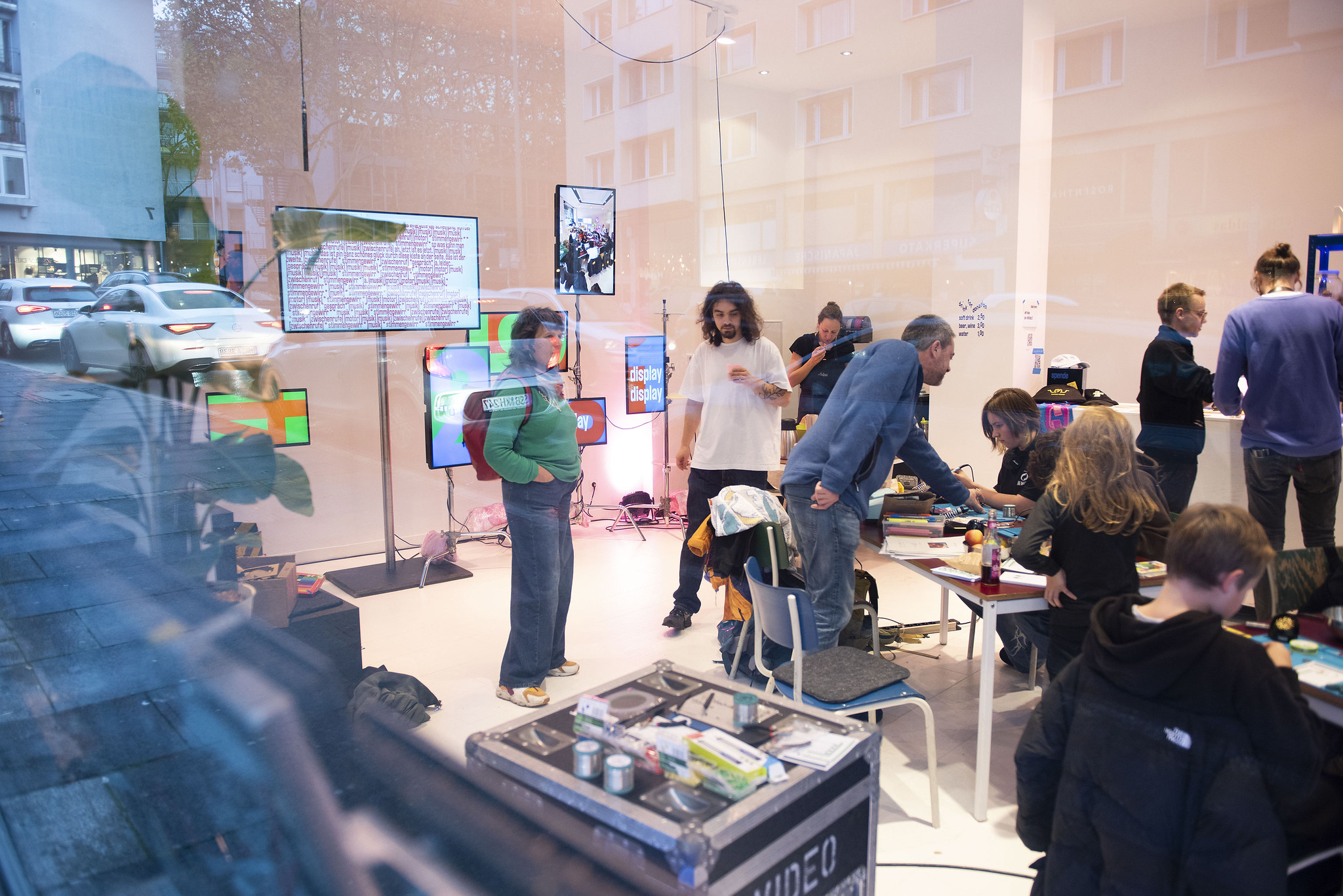 Shot through a window, we see people in a crowded space, in front of an arrangement of colorful digital screens